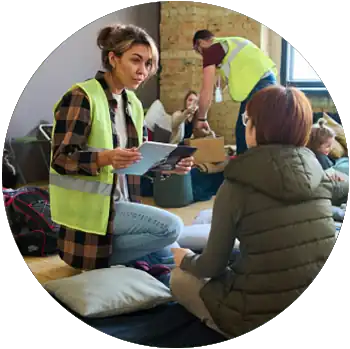 A tan female volunteer with a yellow safety vest holds a tablet and speaks to a person at a shelter.